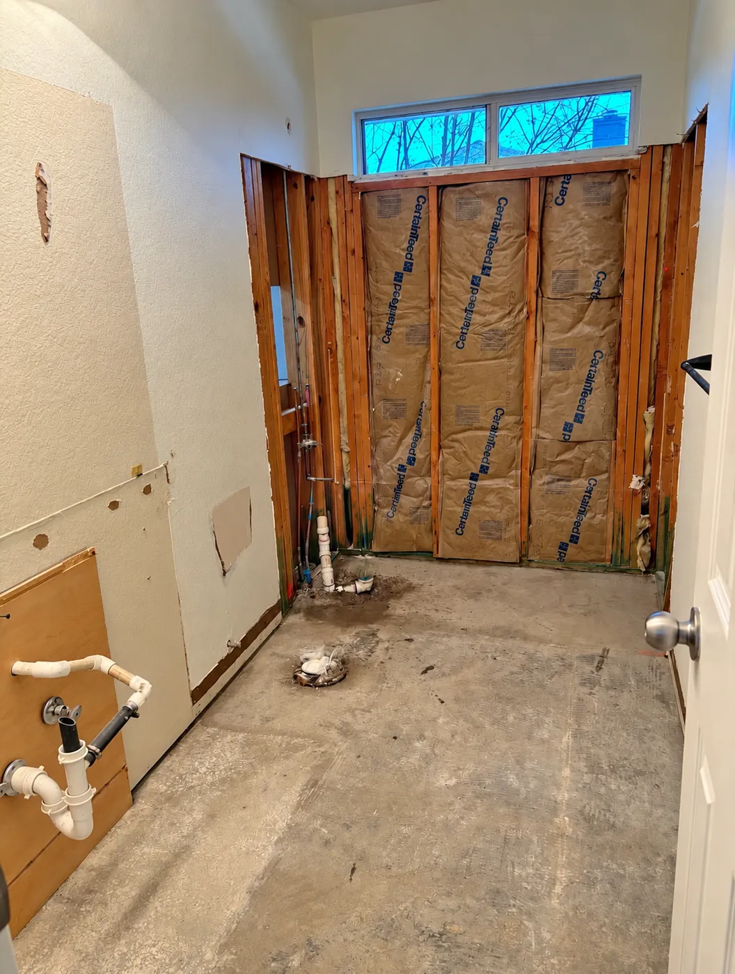 Bathroom during demolition with the tub surround and flooring removed, exposed wall studs, and plumbing stubbed out of the walls