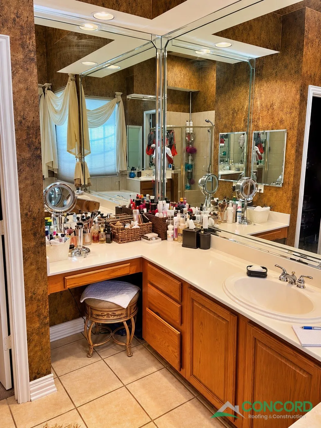 Master bathroom in Frisco, TX before the remodel, with a corner wood vanity and makeup seat, large mirrors, and brown textured walls