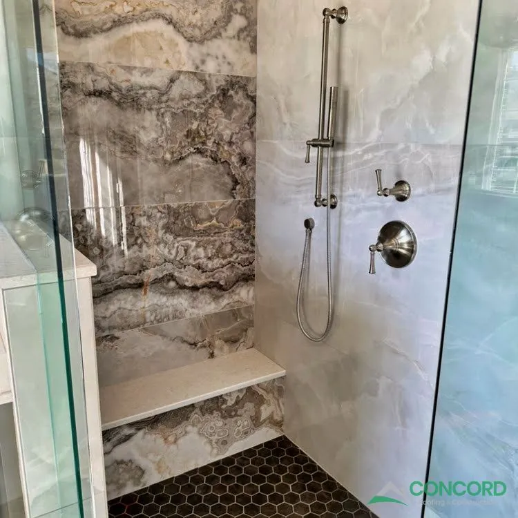 Inside the remodeled shower in a Frisco, TX master bathroom, with large tile, a bench, a handheld shower, and a dark hexagon tile floor
