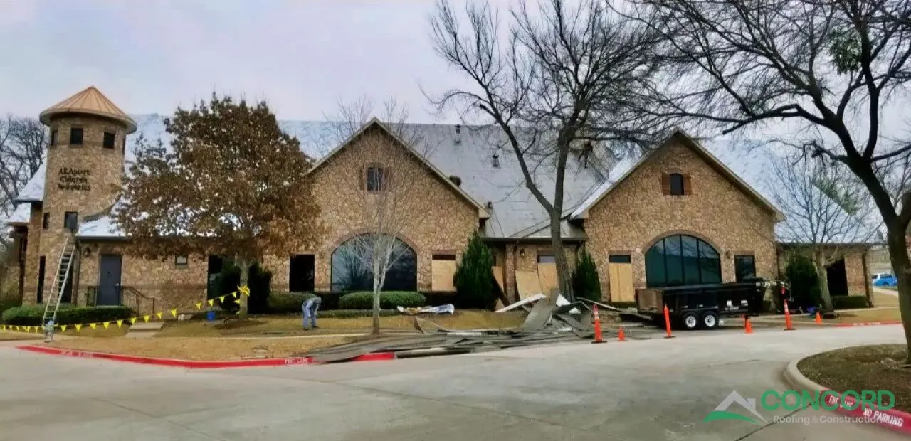 Old roof being torn off a stone pediatric clinic building in McKinney, TX, with plywood over the windows and a trailer for debris
