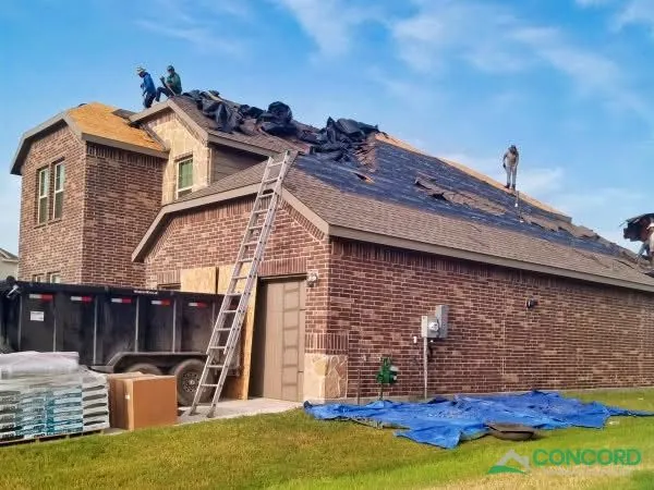 Roofing crew tearing off storm-damaged shingles on a two-story brick home in Princeton, TX, with plywood over the garage door and a dumpster below